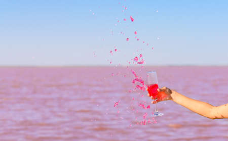 Small splashes of rose wine splashed out of the glass by a girl. In the background there is a beautiful pink water of a salt lake. Selective focusing.の写真素材
