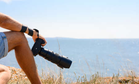 Close-up of a camera in a man's hand on the edge of a steep cliff over the sea. The traveler photographer sits on the edge of the mountain.の写真素材