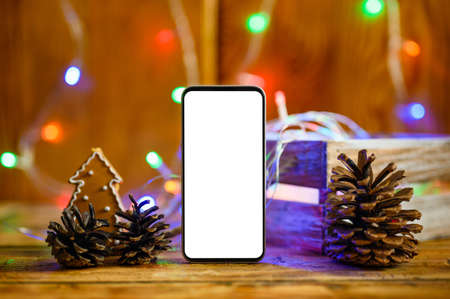 A smartphone with a blank white display stands next to pine cones. In the background there are bokeh from colorful garlands and Christmas decorations. Selective focusing.の写真素材