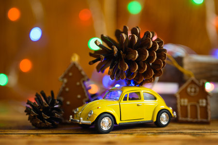 Vladimir, Russia-October 2021: Small toy yellow car with a big beautiful Christmas tree cone on the roof against the background of Christmas lights. Christmas preparation concept. Selective focus on the car.のeditorial素材