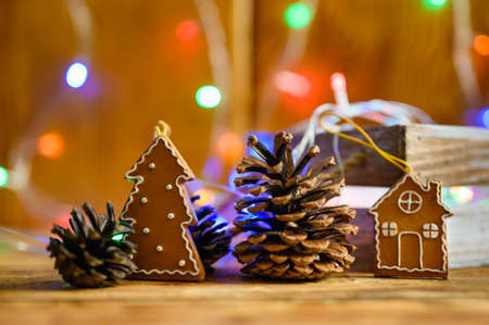 Greeting Christmas card in rustic style. Toy cookies in the shape of a Christmas tree and a house next to pine cones on a wooden background and colorful lights. Selective focus on the tree.の写真素材
