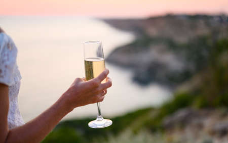 A glass of champagne in the girl's hand close-up on the background of the sunset and the sea. Selective focus on the glass.の写真素材