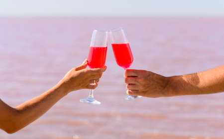 A newlywed couple on honeymoon. Glasses with a pink cocktail of a man and a woman in their hands against the background of a pink lake. Selective focus on a musky hand holding a glassの写真素材