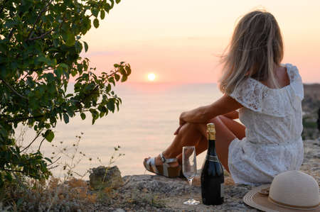 The girl sits alone on the edge of a cliff with a bottle of champagne and a glass. Sunset on the mountain overlooking the sea. View from the back. Selective focus.の写真素材