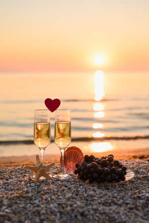 A romantic evening at the sea for a couple in love. A small heart between two glasses of champagne. Nearby seashells and grapes. Selective focus on glass, vertical formatの写真素材