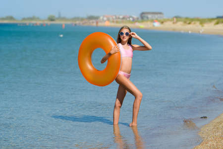 A teenage girl in a pink swimsuit with an orange rubber ring stands on the beach. Portrait of a girl...