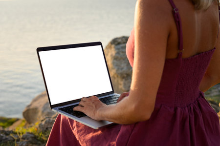A girl in the evening sits by the sea with a computer. Computer with blank white display.の写真素材