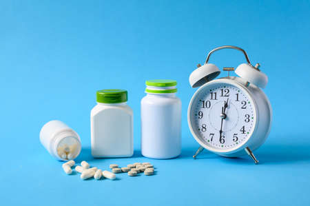 Tablets and pills with cans of medicines next to an alarm clock on a blue background. The concept of taking medications on a schedule.の写真素材