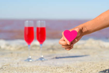 A small pink heart in a woman's hand against the background of two glasses with pink champagne and a pink lake. The end of a romantic meeting. Selective focus on the heart.の写真素材