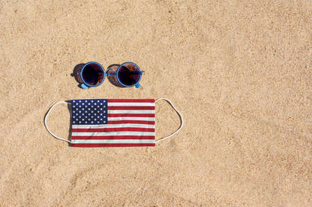 A medical mask in the color of the USA flag lies on the sandy beach next to the glasses. The concept of organizing a safe beach holiday in the United States during the virus pandemic.の写真素材
