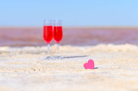 Small pink heart on the background of glasses with rose wine and pink lake. Romantic getaway concept. Selective focus on the heart.の写真素材