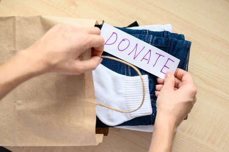 Close-up of a girl lays a stack of donation clothes in a kraft paper bag. Selective focus on donation lettering.の写真素材