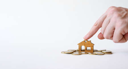 A man's hand points to a figurine of a wooden house and a pile of coins on a white background. Real estate purchase concept. banner, copyspace.の写真素材
