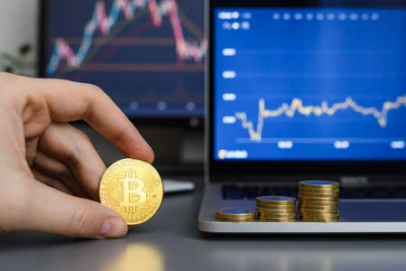 A bitcoin coin in a man's hand against the background of stacks of coins and growing income graphs on monitors. Selective focus on bitcoin.の写真素材