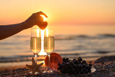 A woman's hand puts a heart on glasses of champagne on the beach against the backdrop of sunset. Preparing for a romantic evening. Selective focus on the hand and on the glass, narrow focus.の写真素材