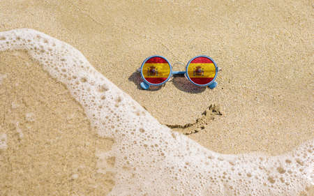 Sunglasses with Spanish flags on a sandy beach. Nearby is a sea lightning and a painted smile. The concept of a successful vacation in the resorts of Spain.の写真素材