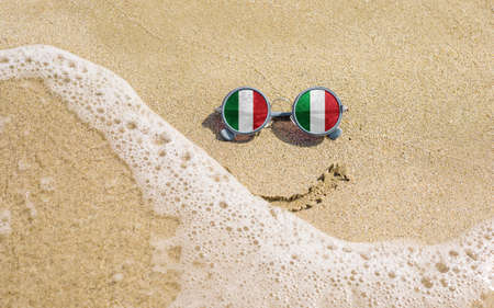 Sunglasses with Italian flags on a sandy beach. Nearby is a sea lightning and a painted smile. The concept of a successful vacation in the resorts of Italy.の写真素材