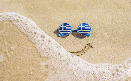 Sunglasses with Greek flags on a sandy beach. Nearby is a sea lightning and a painted smile. The concept of a successful vacation in the resorts of Greece.の写真素材