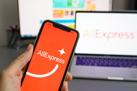 Vladimir, Russia - January 2022: Smartphone with AliExpress application in hand on the background of a laptop. The concept of online shopping for goods from China. Selective focus on the logo.のeditorial素材
