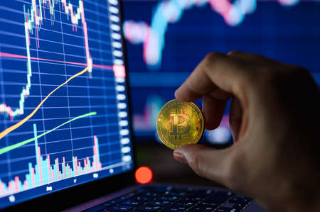 Bitcoin coin in hand next to financial charts on monitors. Selective focus on bitcoin.の写真素材