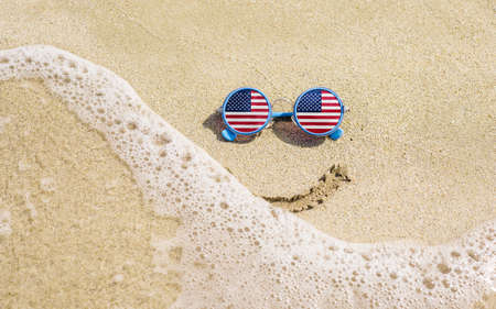 Sunglasses with flag of USA on a sandy beach. Nearby is a sea lightning and a painted smile. The concept of a successful vacation in the resorts of USA.の写真素材