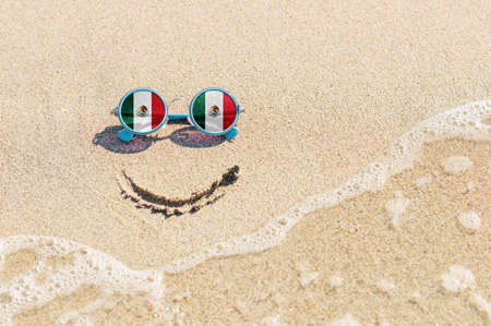 A painted smile on the sand and sunglasses with the flag of Mexico. The concept of a positive and successful holiday in the resort of Mexico.の写真素材