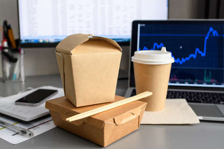 The concept of food delivery to the office to the workplace. Paper containers for food and wooden chopsticks on the table against the background of a laptop and a computer. selective focus.の写真素材