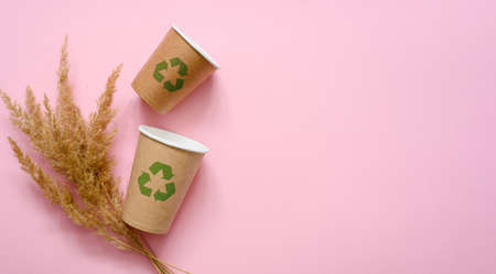 Two paper eco-friendly disposable cups with a green recycling symbol on a pink background. Nearby is a bouquet of dry grass. The concept of using biodegradable materials to protect the environment.の写真素材
