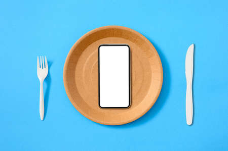 A smartphone with a blank white display on a brown paper empty plate on a blue background. Food delivery service advertising concept. flat lay.の写真素材