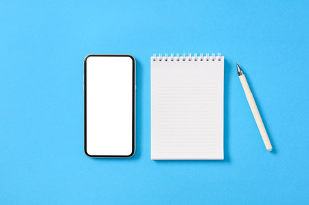Smartphone with a blank white display and a notepad with a pen on a blue paper background. Flat lay, mock up.の写真素材