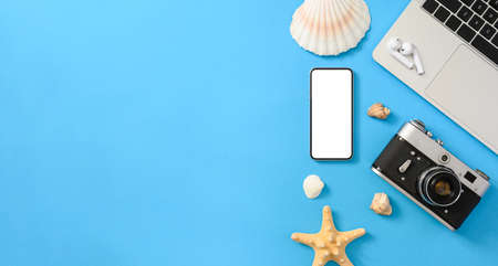 Top view of blank screen smartphone, camera, laptop and some seashells on blue paper background. Travel preparation concept. Flat lay, copy space, mock up.の写真素材