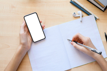 A smartphone with an isolated white display in the hand of a schoolboy against the background of a desk with a notebook. Selective focus on smartphone, top view.の写真素材