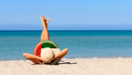 A slender tanned girl on the beach in a straw hat in the colors of the flag of UAE. The concept of a perfect vacation in a resort in the United Arab Emirates. Focus on the hat.の写真素材
