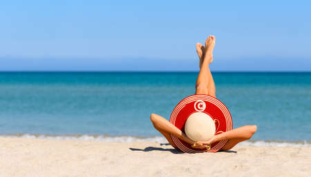 A slender girl on the beach in a straw hat in the colors of the Tunisia flag. The concept of a perfect vacation in a resort in the Tunisia. Focus on the hat.の写真素材