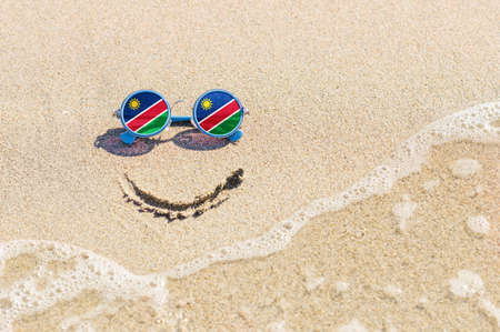 A painted smile on the sand and sunglasses with the flag of the Namibia. The concept of a positive and successful holiday in the resort of the Namibia.の写真素材