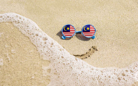 Sunglasses with flag of Malaysia on a sandy beach. Nearby is a sea lightning and a painted smile. The concept of a successful vacation in the resorts of the Malaysia.の写真素材