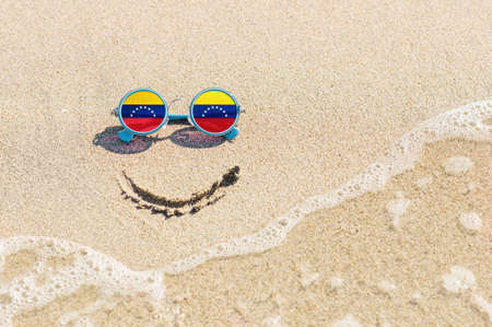 A painted smile on the sand and sunglasses with the flag of Venezuela. The concept of a positive and successful holiday in the resort of Venezuela.の写真素材