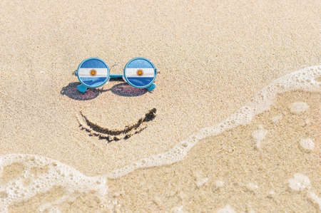 A painted smile on the sand and sunglasses with the flag of Argentina. The concept of a positive and successful holiday in the resort of Argentina.の写真素材