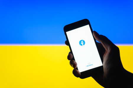 Vladimir, Russia - April 2022: Starting the Facebook app on a smartphone against the background of the Ukrainian flag. Selective focus on logoのeditorial素材