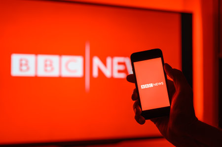 Vladimir, Russia - February 2022: Smartphone in hand with BBS application for viewing world news on the background of the BBC channel. Selective focus on the logo.のeditorial素材