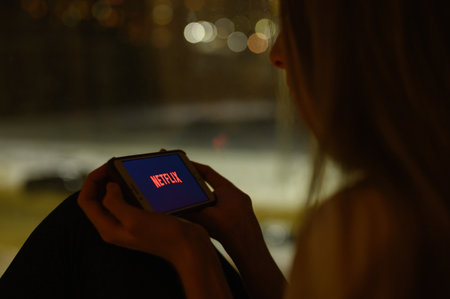 Vladimir, Russia - January 2022: A girl at night on a window on a smartphone opens an application for watching Netflix series. Selective focus on the logo.のeditorial素材