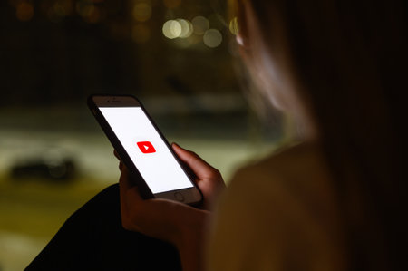 Vladimir, Russia - January 2022: YouTube application on a smartphone in the hands of a girl at night. Social media addiction concept. Selective focus on the logo, narrow focus.のeditorial素材