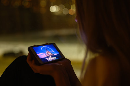 Vladimir, Russia - January 2022: Girl watching a movie produced by Disney on a smartphone at night....