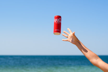 Anapa, Russia - July 2021: Girl on the beach catches a can of Coca-Cola against the background of the sea and sky. Close-up of hand and flying drink canのeditorial素材