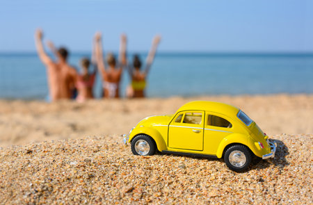 Anapa, Russia - August 2021: A toy car stands on the beach against the background of the sea and a sitting happy family. Selective focus on the car.のeditorial素材