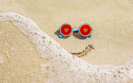 Sunglasses with flag of Montenegro on a sandy beach. Nearby is a sea lightning and a painted smile. The concept of a successful vacation in the resorts of Montenegro.の写真素材