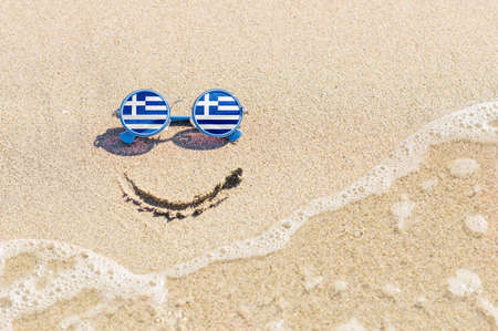 A painted smile on the beach and sunglasses with the flag of Greece. The concept of a positive holiday in the resort of Greece.の写真素材
