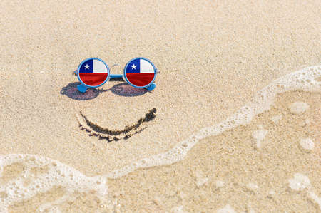A painted smile on the beach and sunglasses with the flag of the Chile. The concept of a positive holiday in the resort of the Chile.の写真素材