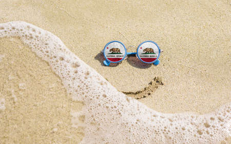 Sunglasses with flag of the State of California on a sandy beach. Nearby is a sea lightning and a painted smile. The concept of a successful vacation in the resorts of California.の写真素材