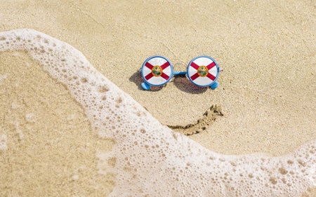 Sunglasses with flag of the State of Florida on a sandy beach. Nearby is a sea lightning and a painted smile. The concept of a successful vacation in the resorts of Florida.の写真素材